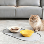 Um gato ao lado de duas tigelas sobre um tapete oval cinza. Tigelas e alimento não incluídos; selecione uma cor disponível do tapete.